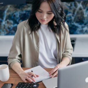 Mujer sentada frente a una computadora portátil, tomando notas en un cuaderno sobre una mesa donde también hay una calculadora, un teléfono celular y una taza. La imagen ilustra el Curso de Búsqueda de Fondos externos.
