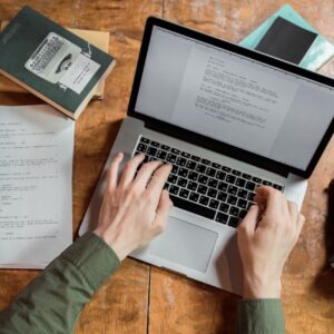 Persona escribiendo en una computadora portátil sobre una mesa de madera, rodeada de documentos impresos, libros y cuadernos. La imagen representa el Curso de Desarrollo de Propuestas.
